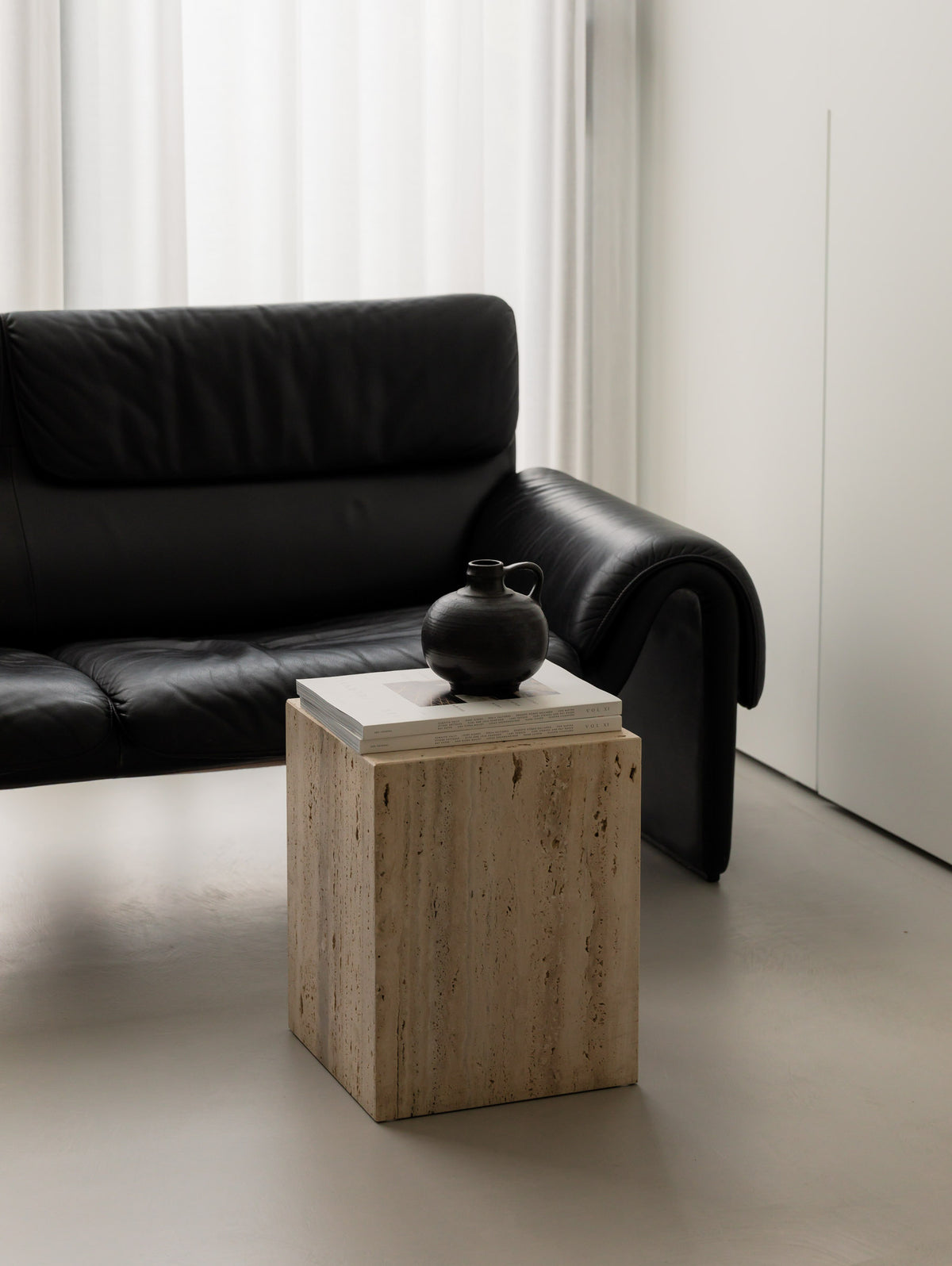 Un'common COI travertine pedestal with a round black ceramic jug on stacked art books, in front of a black leather lounge chair and sheer curtains.