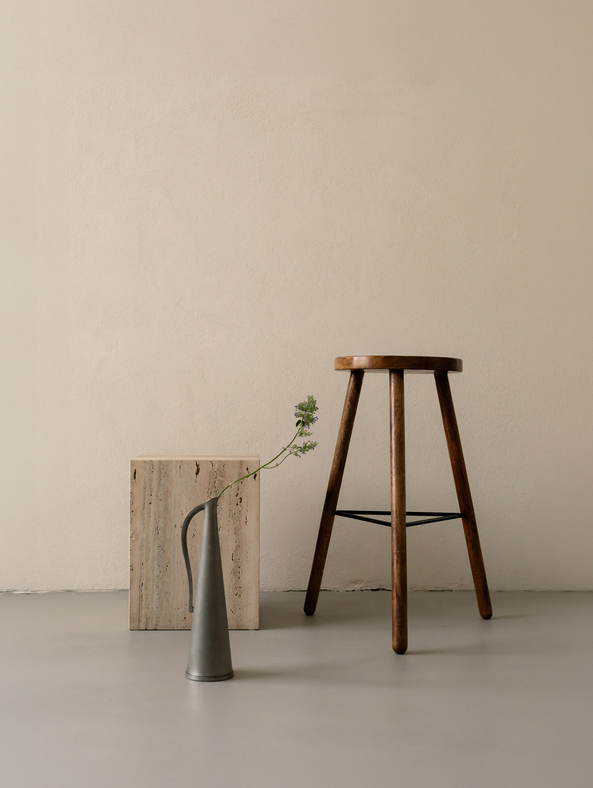 Un'common COI travertine pedestal styled with a pewter jug vase and a single wildflower stem, beside a dark wood tripod stool.
