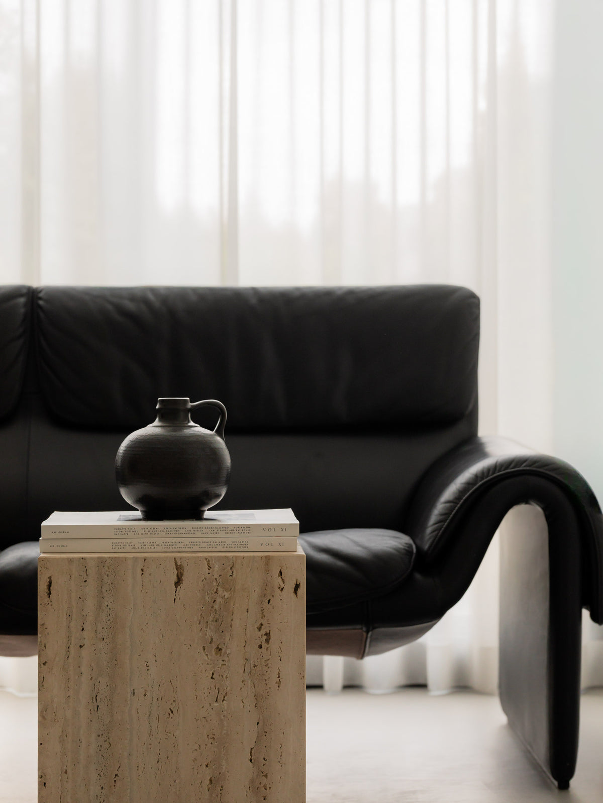 Close-up of a black ceramic jug on stacked art books on Un'common COI travertine pedestal, in front of a black leather lounge sofa.