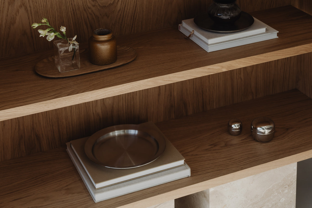 Un'common Sideboard unit in warm oak, styled with a glass bud vase, a stoneware jar, a wooden tray, stacked books, a ceramic lamp base, a steel plate, and small metallic trinket boxes.